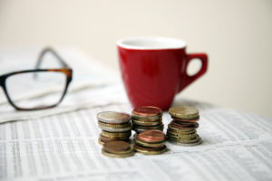Das Bild zeigt aufgestapelte Münzen vor dem Hintergrund einer Kaffeetasse und einer Brille.